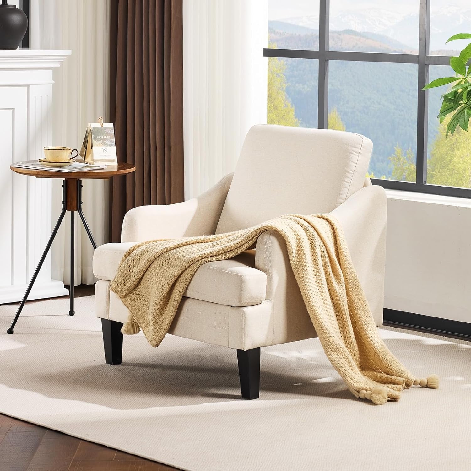 Beige upholstered armchair with a textured tan throw draped over it beside a round side table and large window