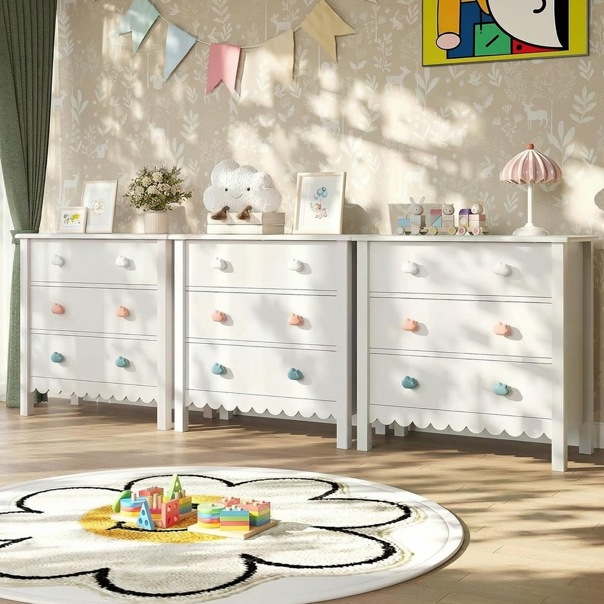 Three white scalloped dressers arranged along a wallpapered wall with toys and decor on top in a playroom.