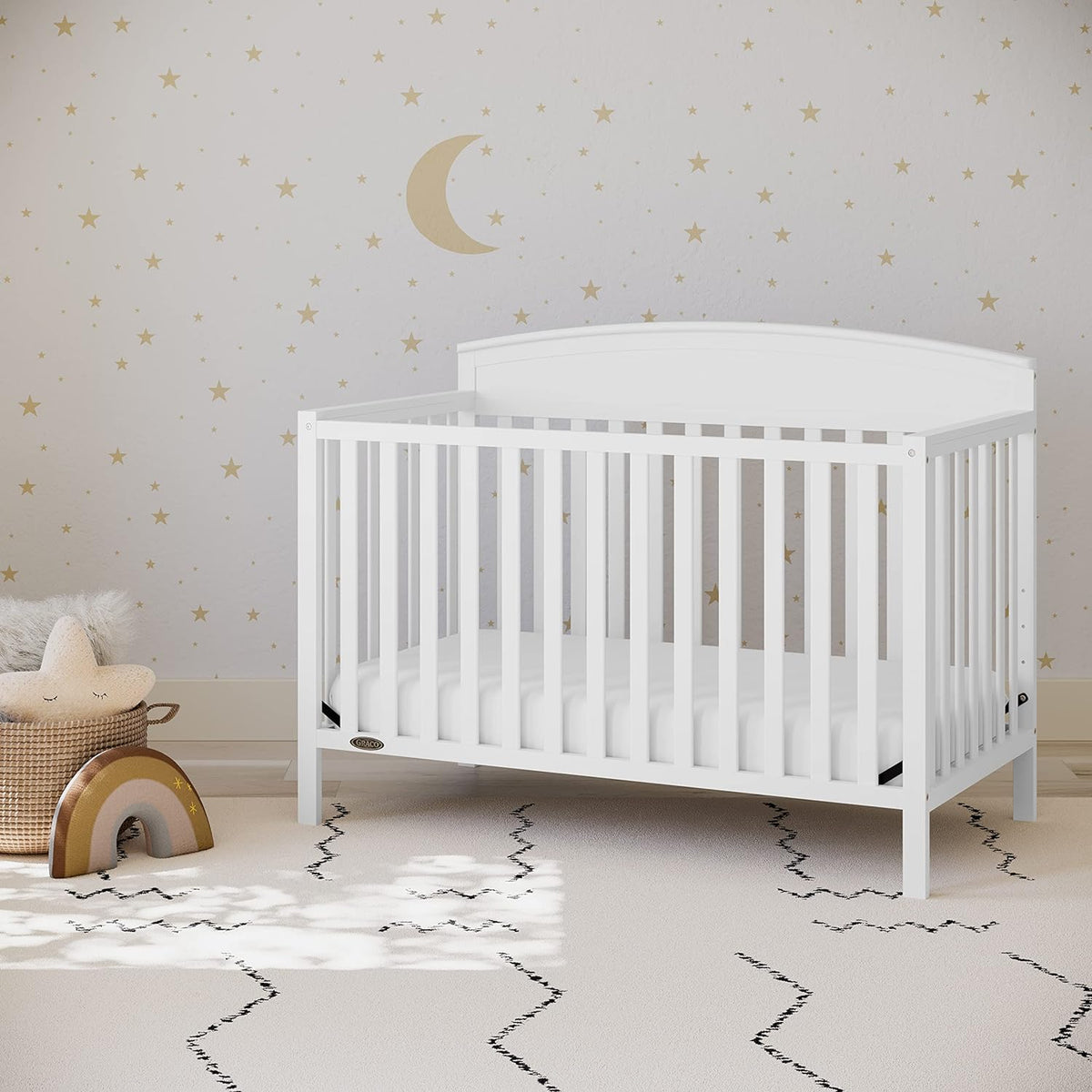 White crib placed in a nursery with star-and-moon wall decals, rug, and a woven basket with toys