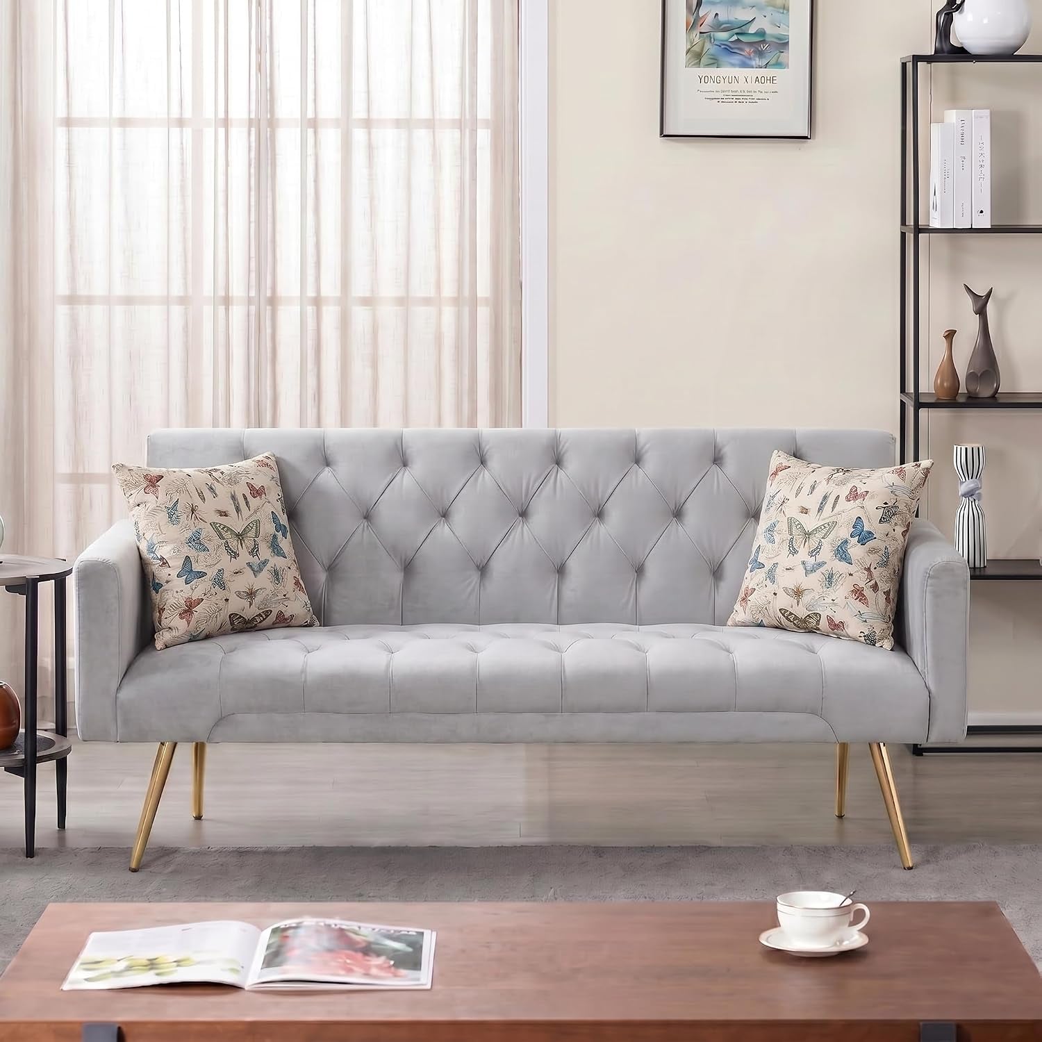Light gray tufted sofa with low arms and two patterned throw pillows, styled against sheer curtains.
