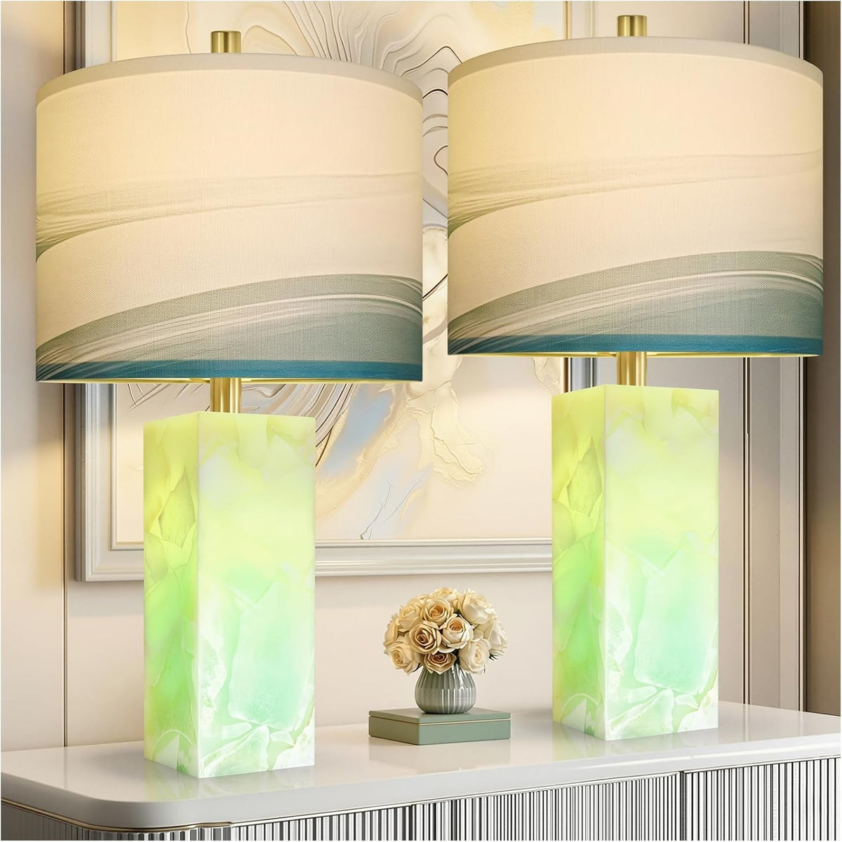 Two glowing pale green alabaster column lamps with cream patterned drum shades on a white console