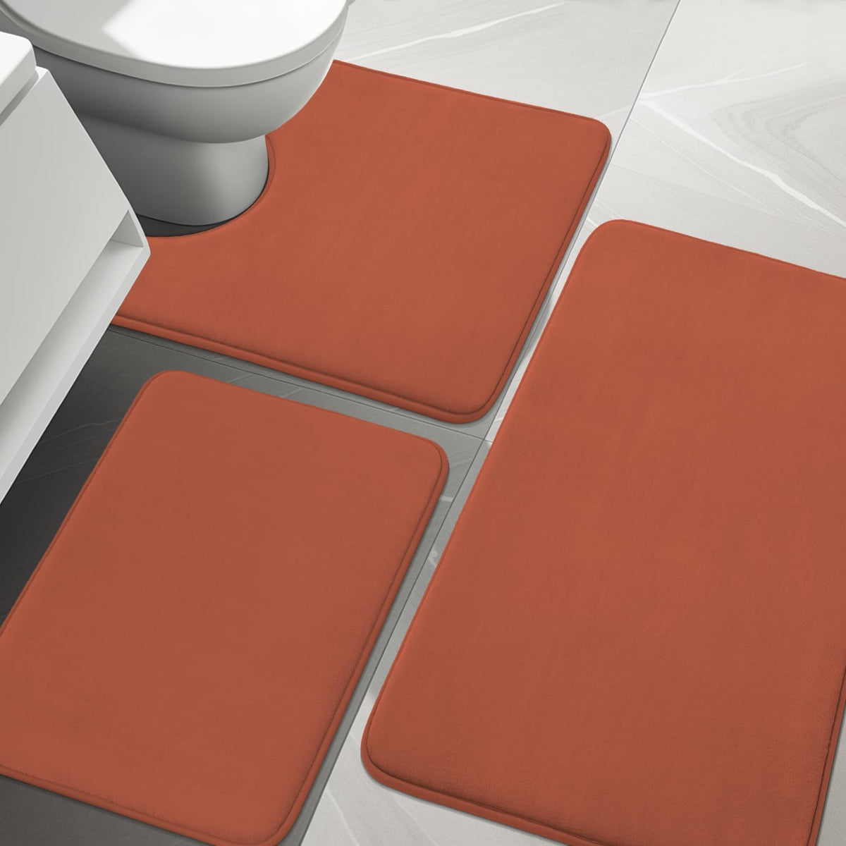 Close-up angled view of three rust-orange memory foam bath rugs on glossy tile.