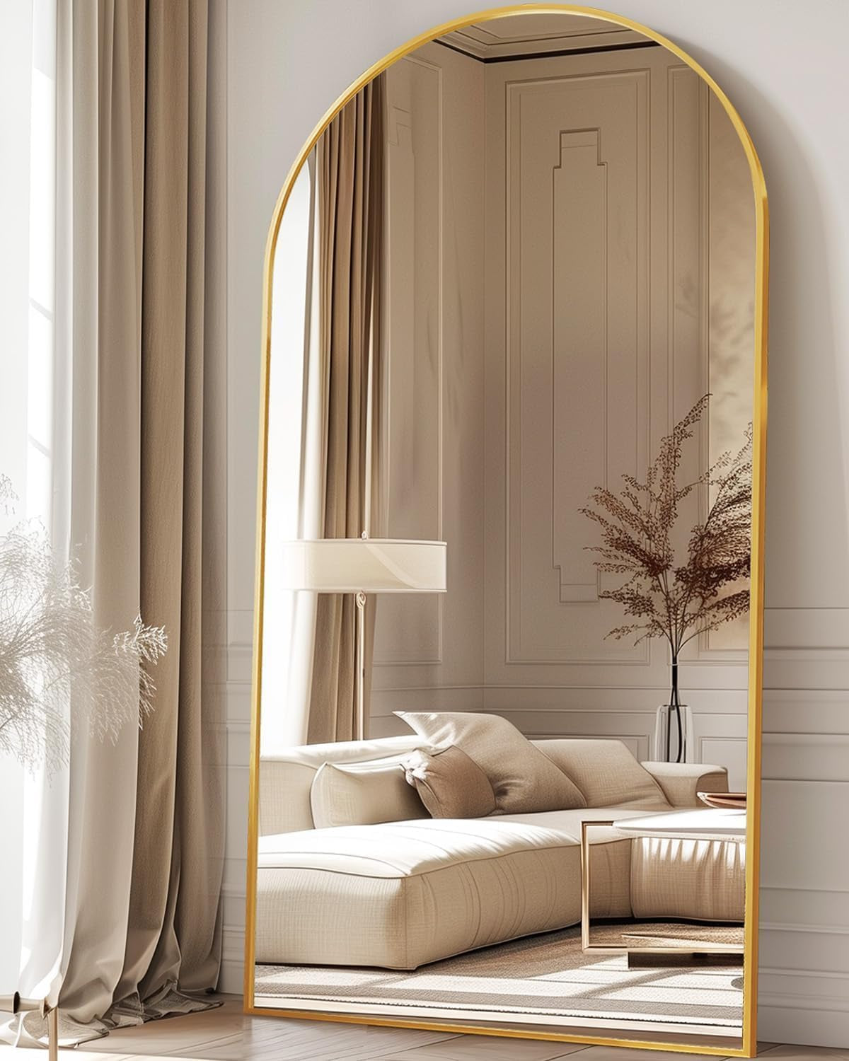 Arched gold-framed floor mirror leaning against a wall, reflecting a beige sofa and drapery.