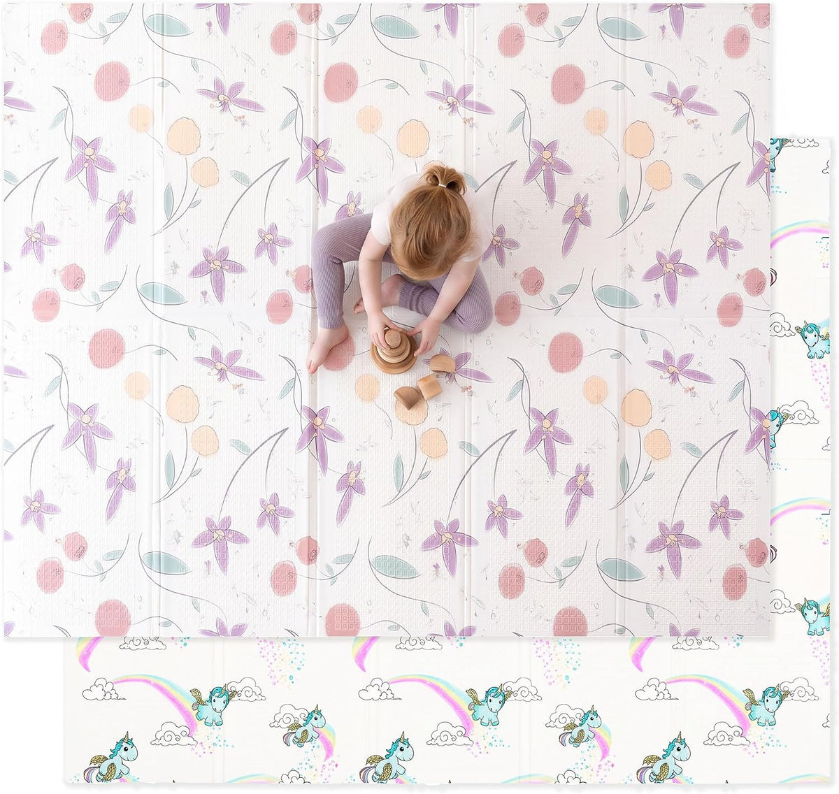 Top-down view of a toddler playing on a light floral mat with purple flowers; a unicorn-and-rainbow print peeks from the