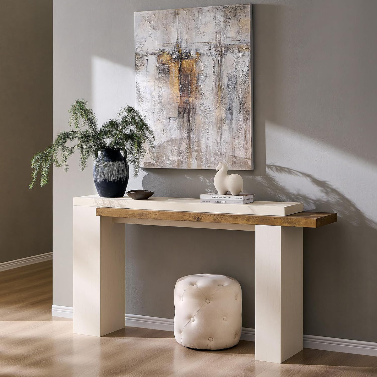 Console table beneath an abstract canvas, topped with a dark ceramic vase and small bowl, tufted round pouf beneath.