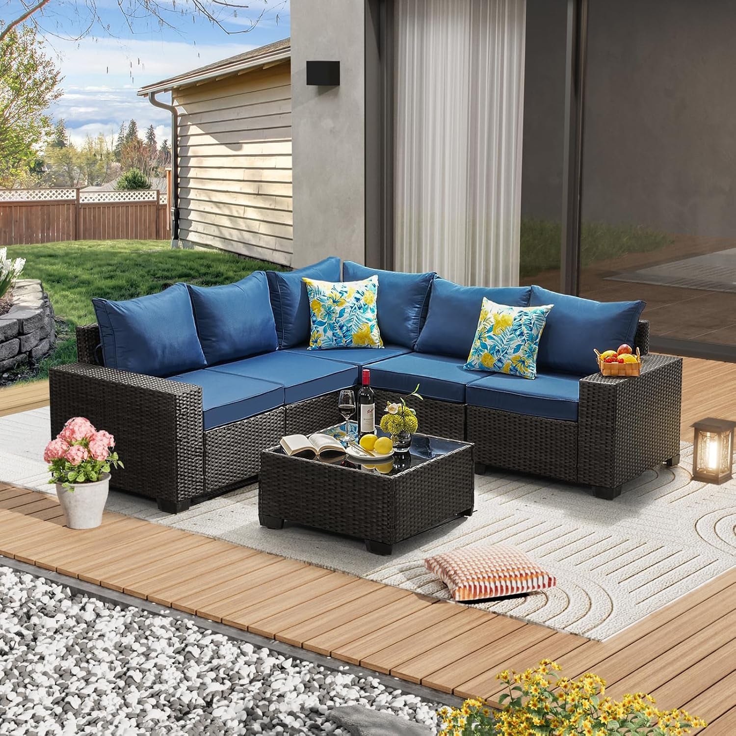 L-shaped rattan patio sofa with bright blue cushions, glass coffee table and decorative pillows on an outdoor rug.