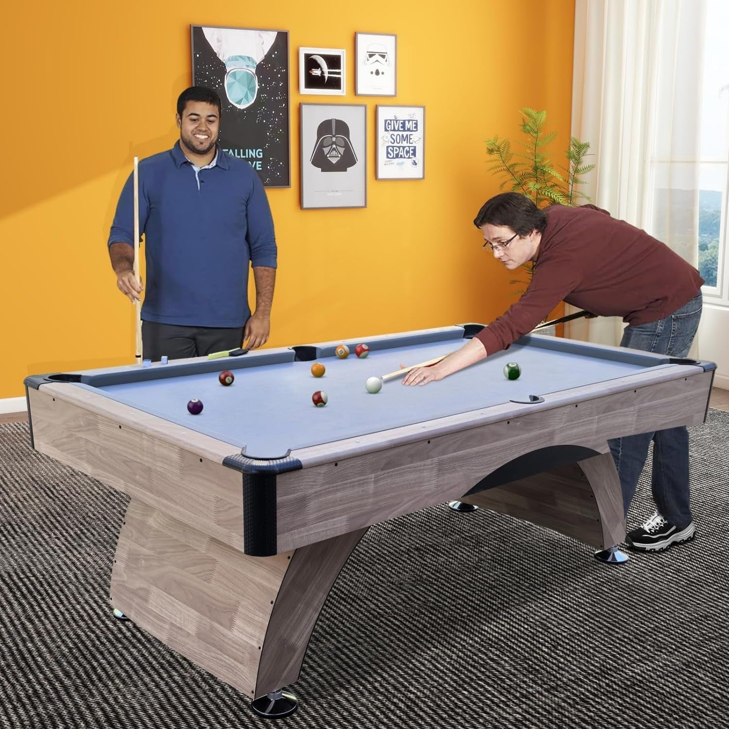 Two men playing on a grey-blue felt 7 ft pool table in a casual game room with an orange accent wall