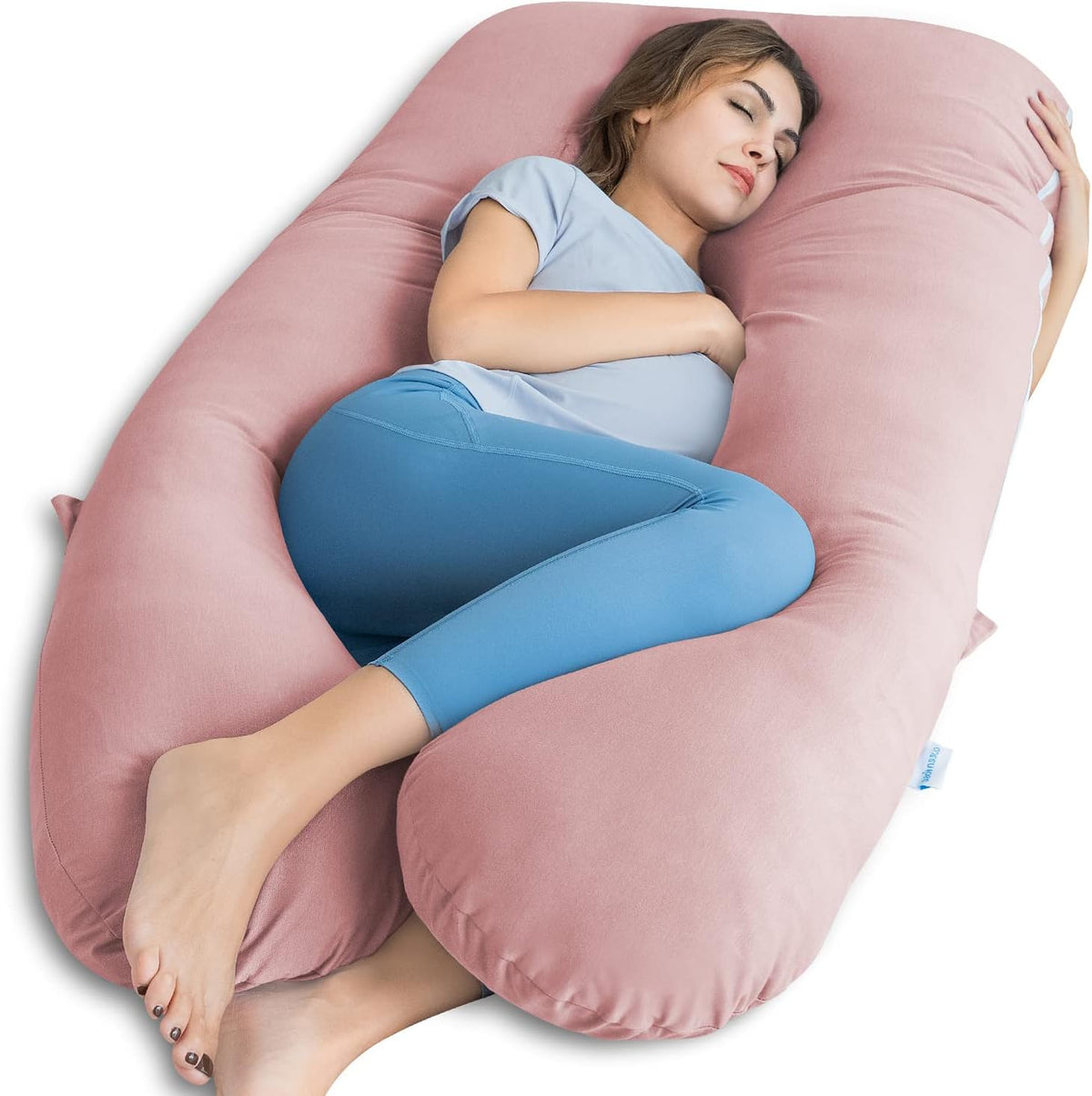 Woman reclining inside a U-shaped blush-pink full-body maternity pillow, curled on her side.