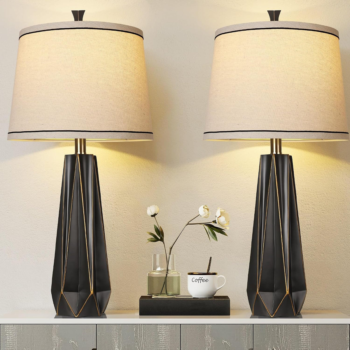 Front view of two black geometric table lamps with beige shades and subtle gold accents beside a vase and coffee cup.