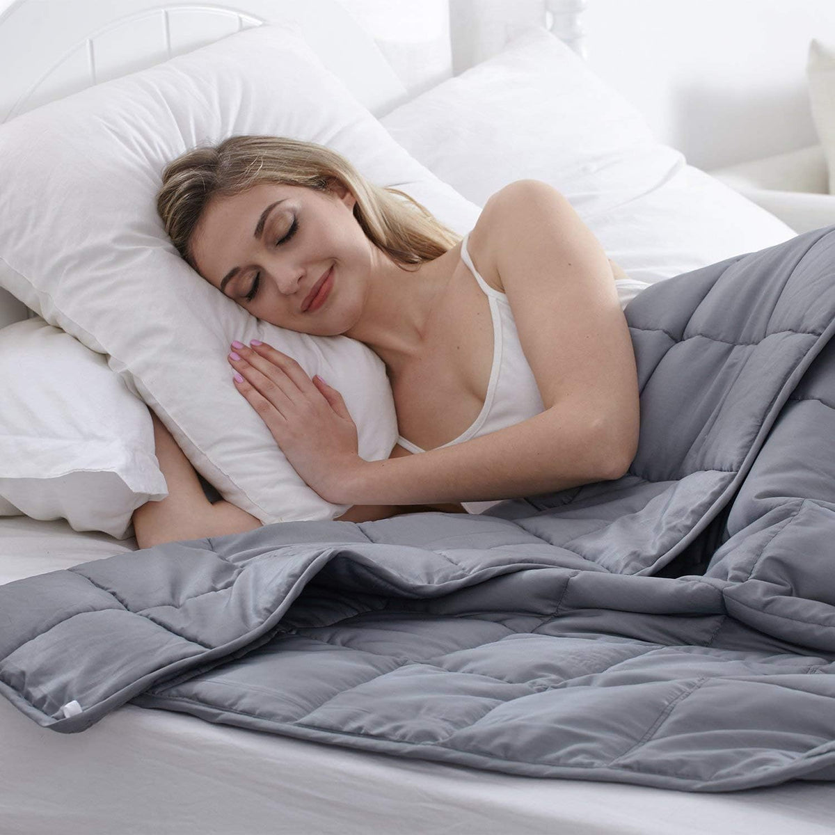 Person sleeping under a dark grey weighted blanket on a bed with white pillows, angled lifestyle view