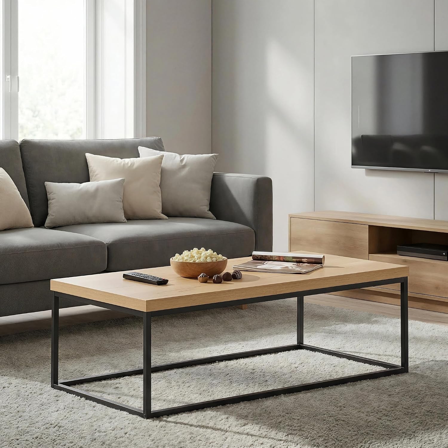 Light wood rectangular coffee table with black metal frame in front of a gray sofa, popcorn bowl and remote on top.