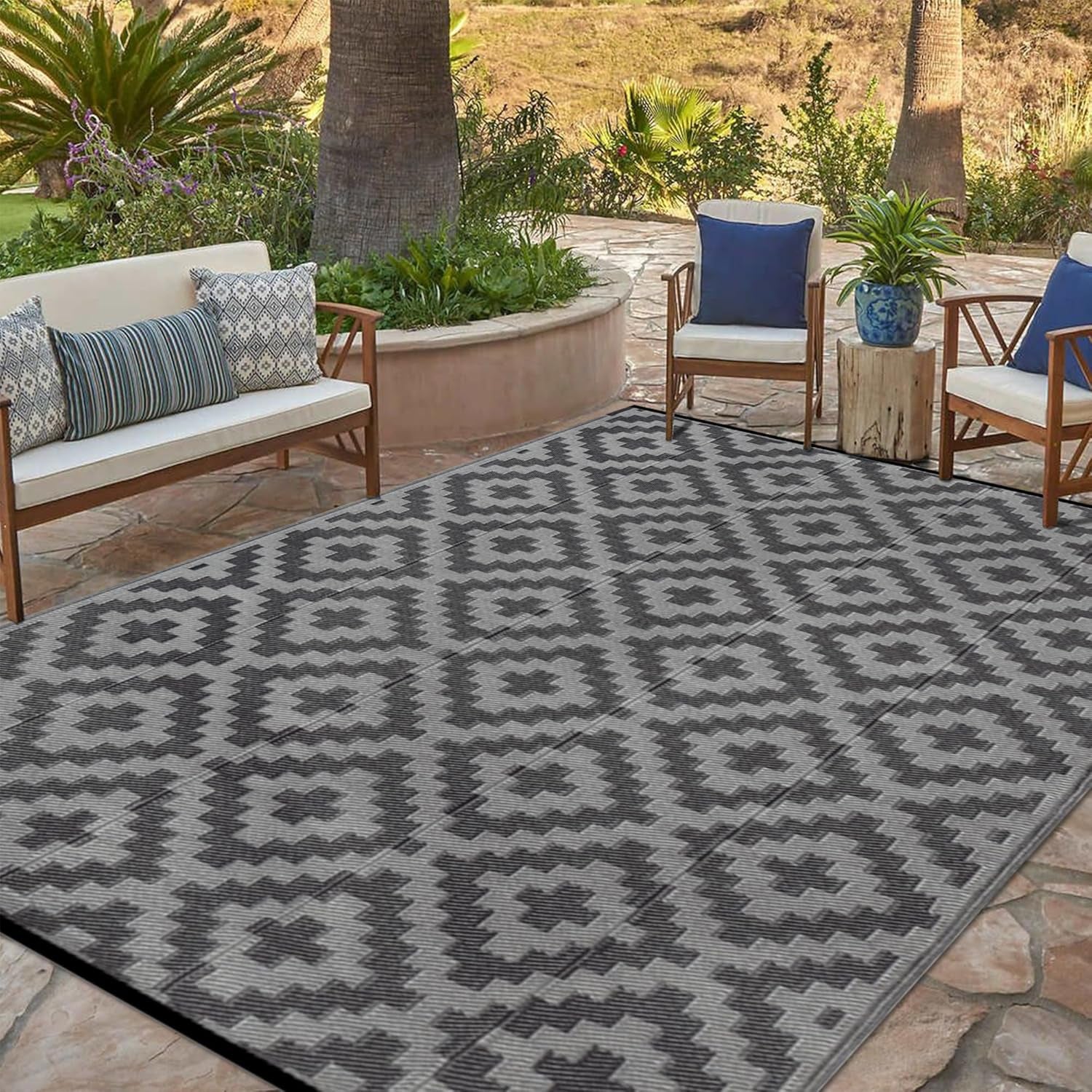 Patio seating with a gray geometric rug beneath a bench and chairs beside potted plants and palm trees.