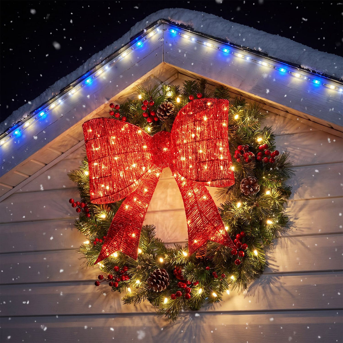 A glowing red bow decorates a green wreath mounted on a snowy roof.