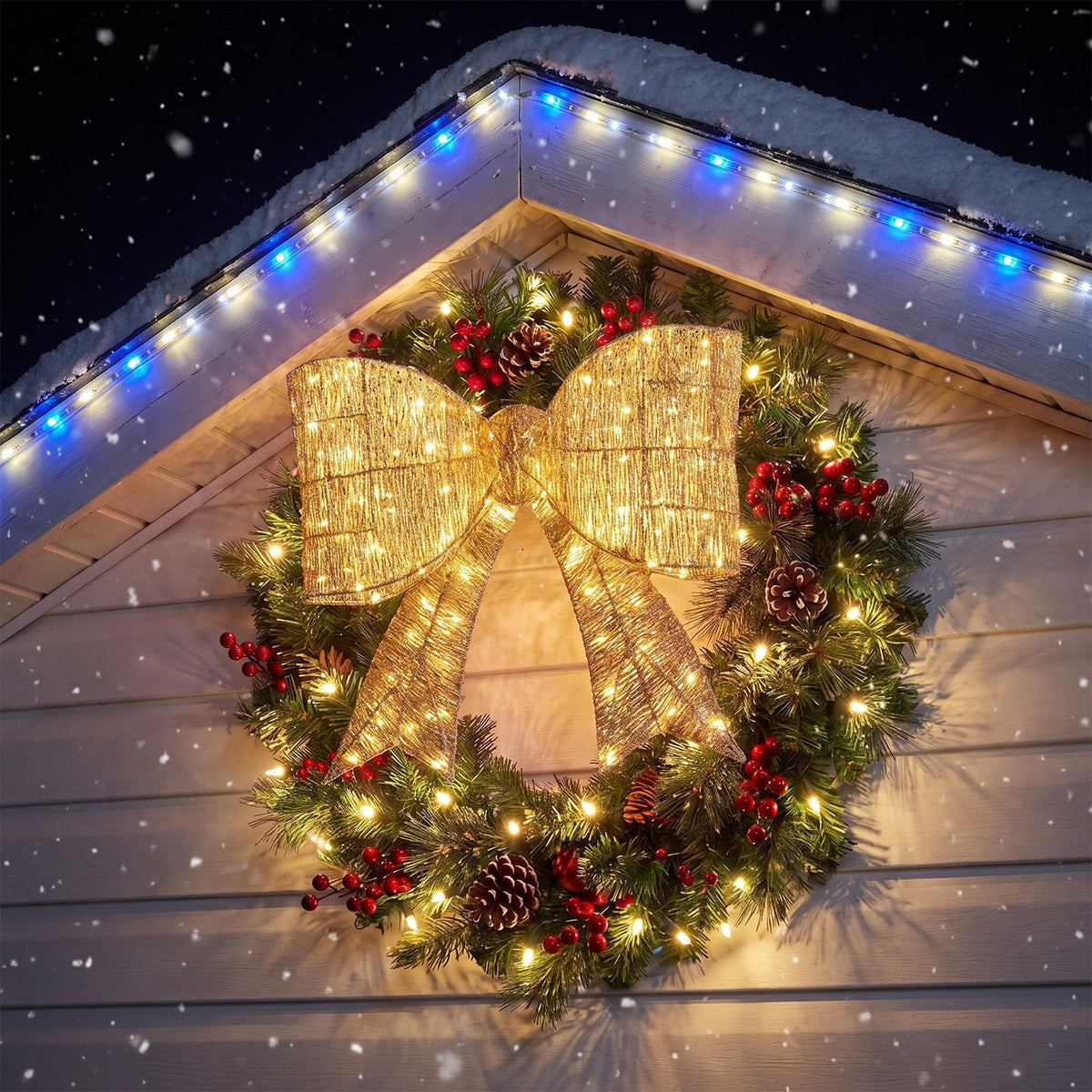 A glowing gold bow decorates a greenery wreath on a snowy house exterior at night.