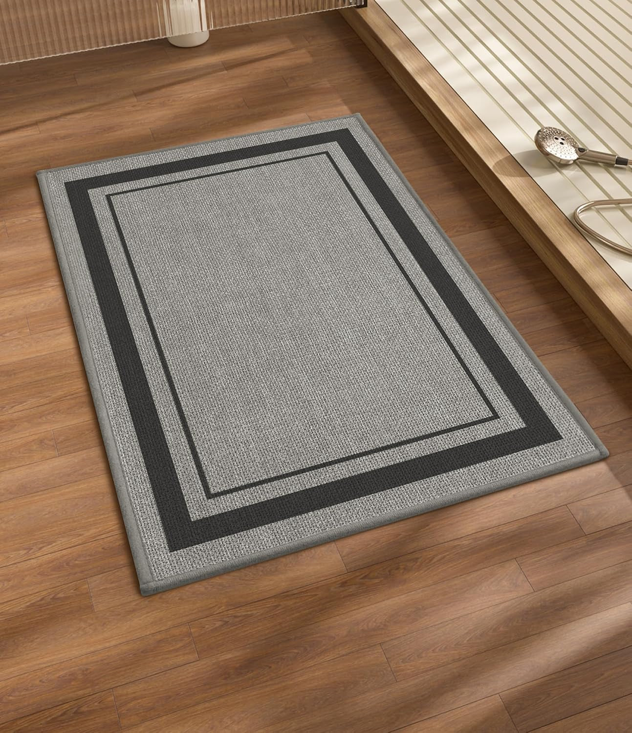 Light gray bath rug with a wide dark gray border lying on wood flooring next to a tub platform.