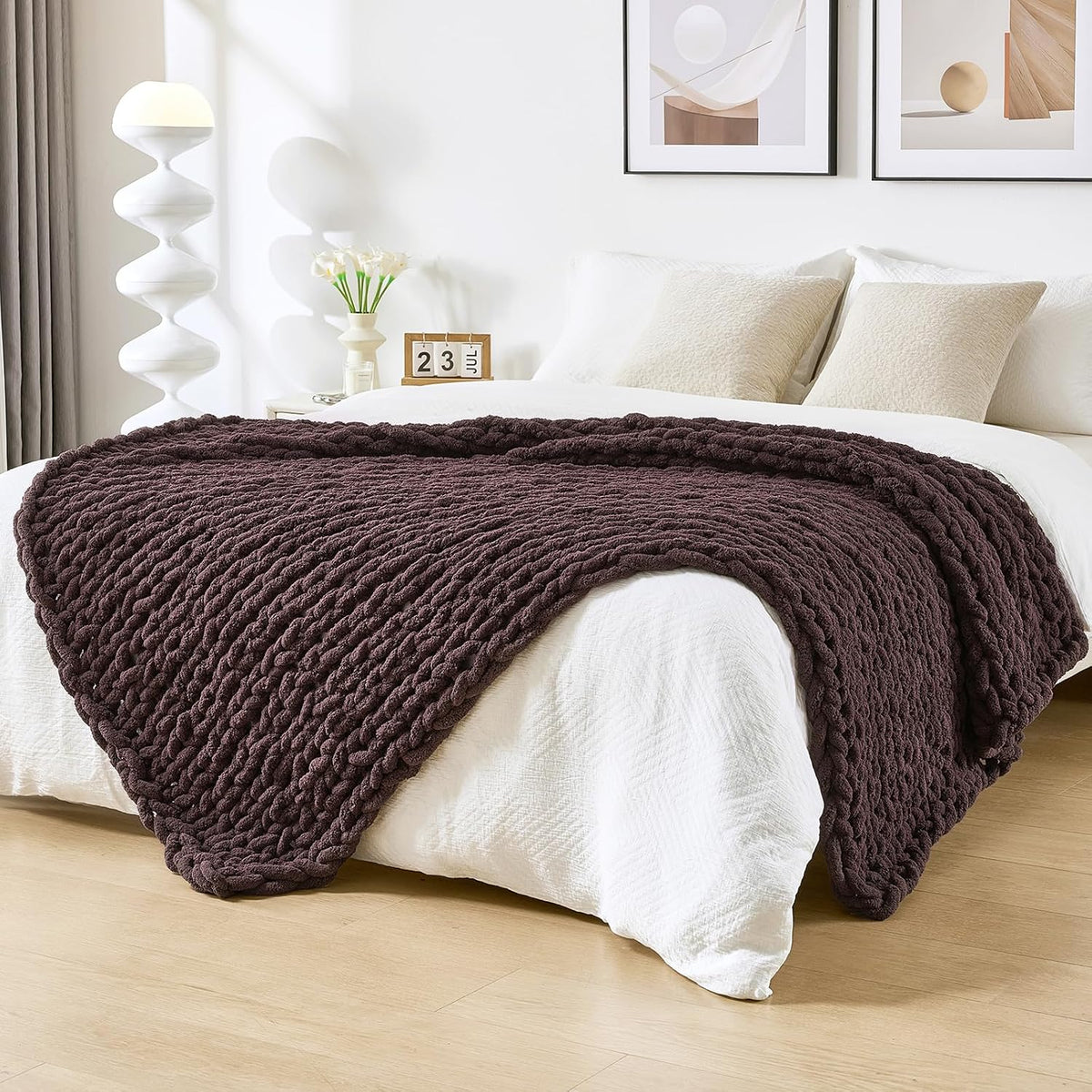 Dark brown chunky knit throw laid across the foot of a white bed in a bright bedroom.