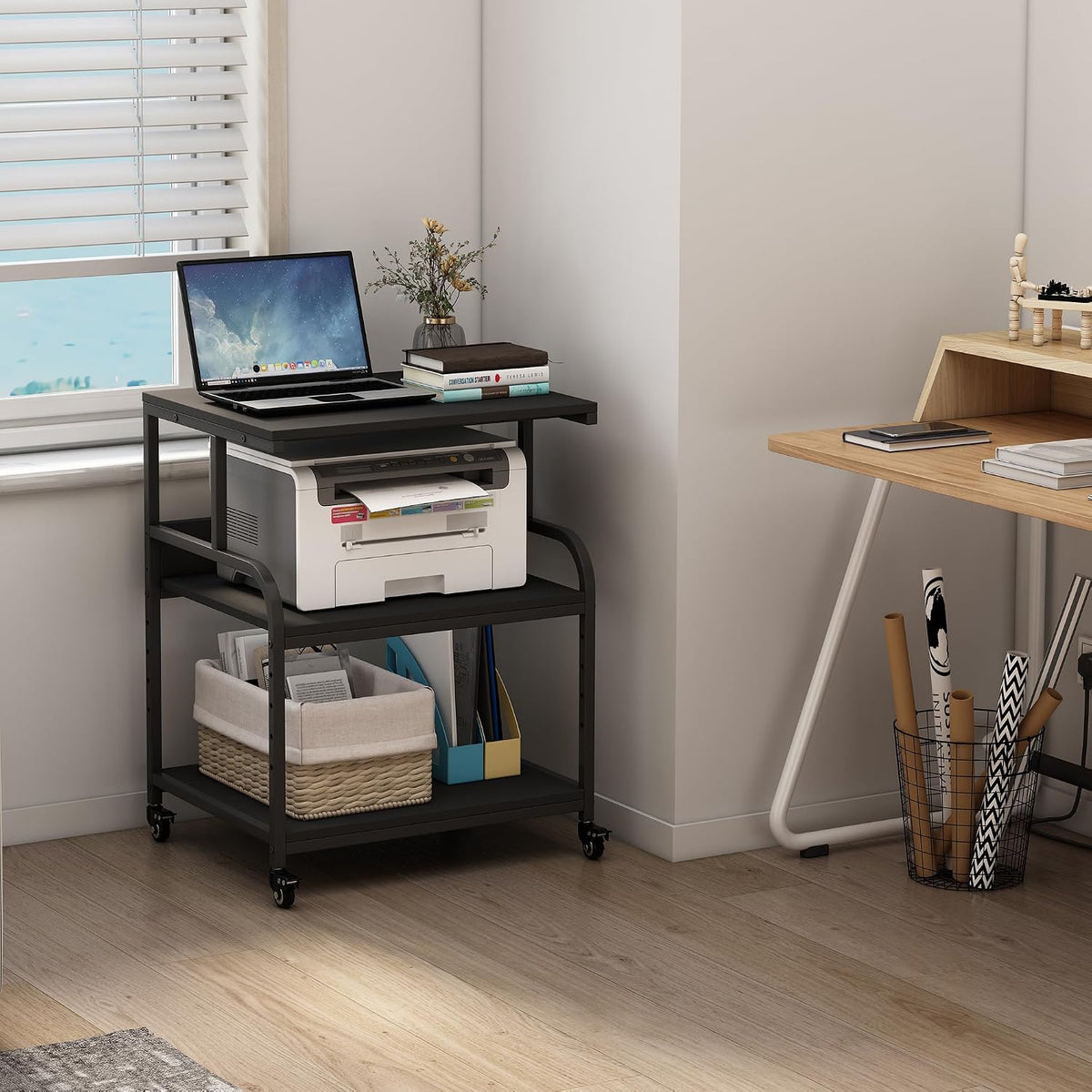 Black three-tier printer stand in a bright nook by a window with a laptop on top and storage basket on the bottom shelf.