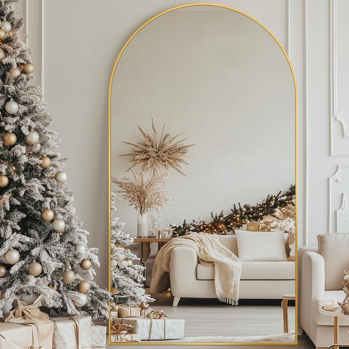 Arched full-length mirror with a thin gold frame standing on the floor beside a decorated Christmas tree.