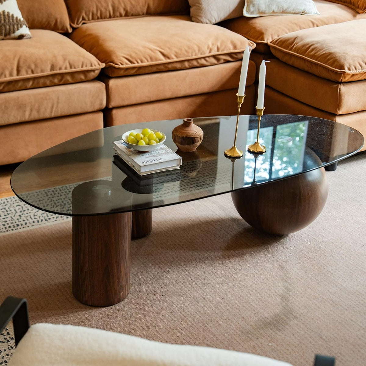 Low oval coffee table with a dark smoked top and walnut‑finish cylindrical and spherical legs before a tan sectional sofa.
