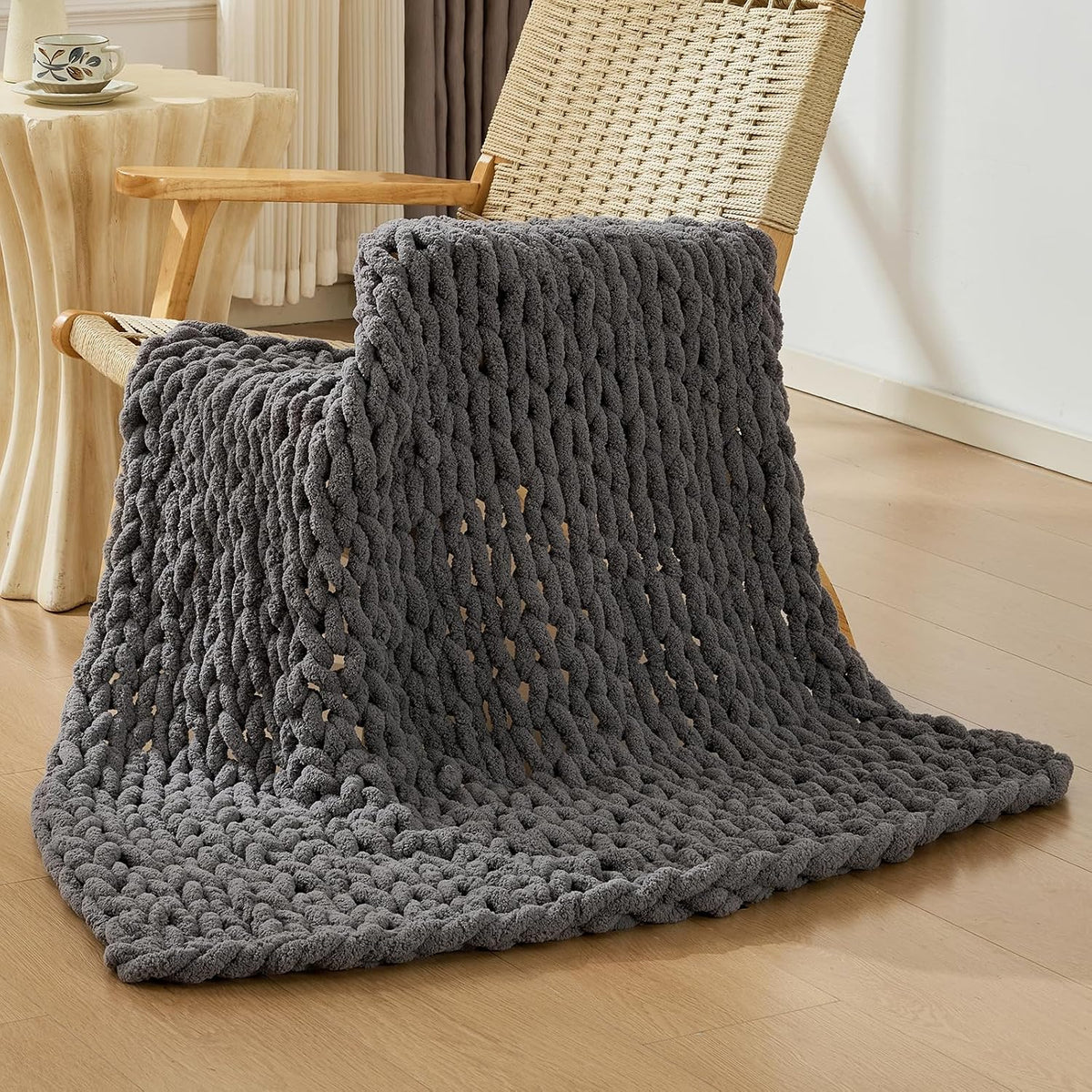 Charcoal chunky knit throw folded over a woven wooden chair arm, pooling on the floor
