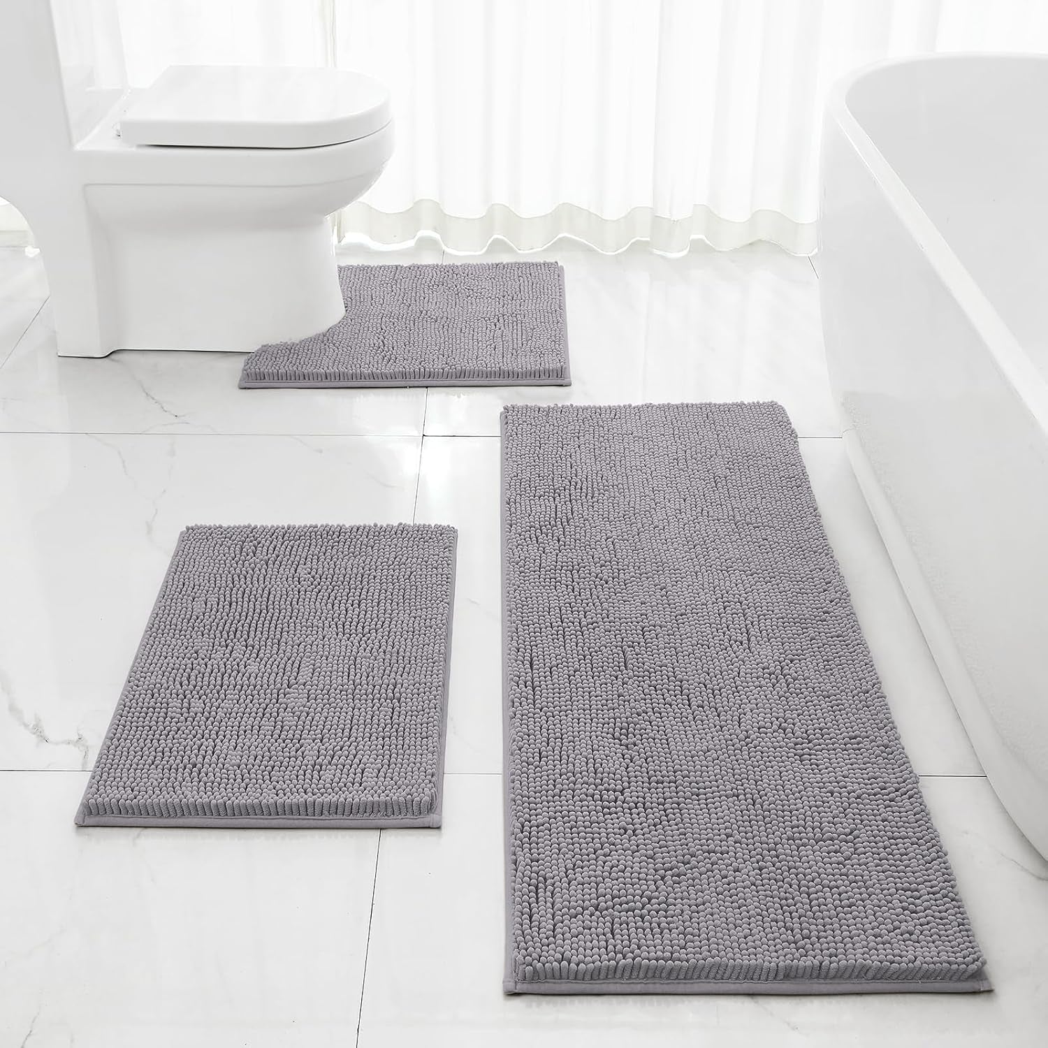 Bathroom scene with three grey chenille mats on a tiled floor near a toilet and a freestanding tub.