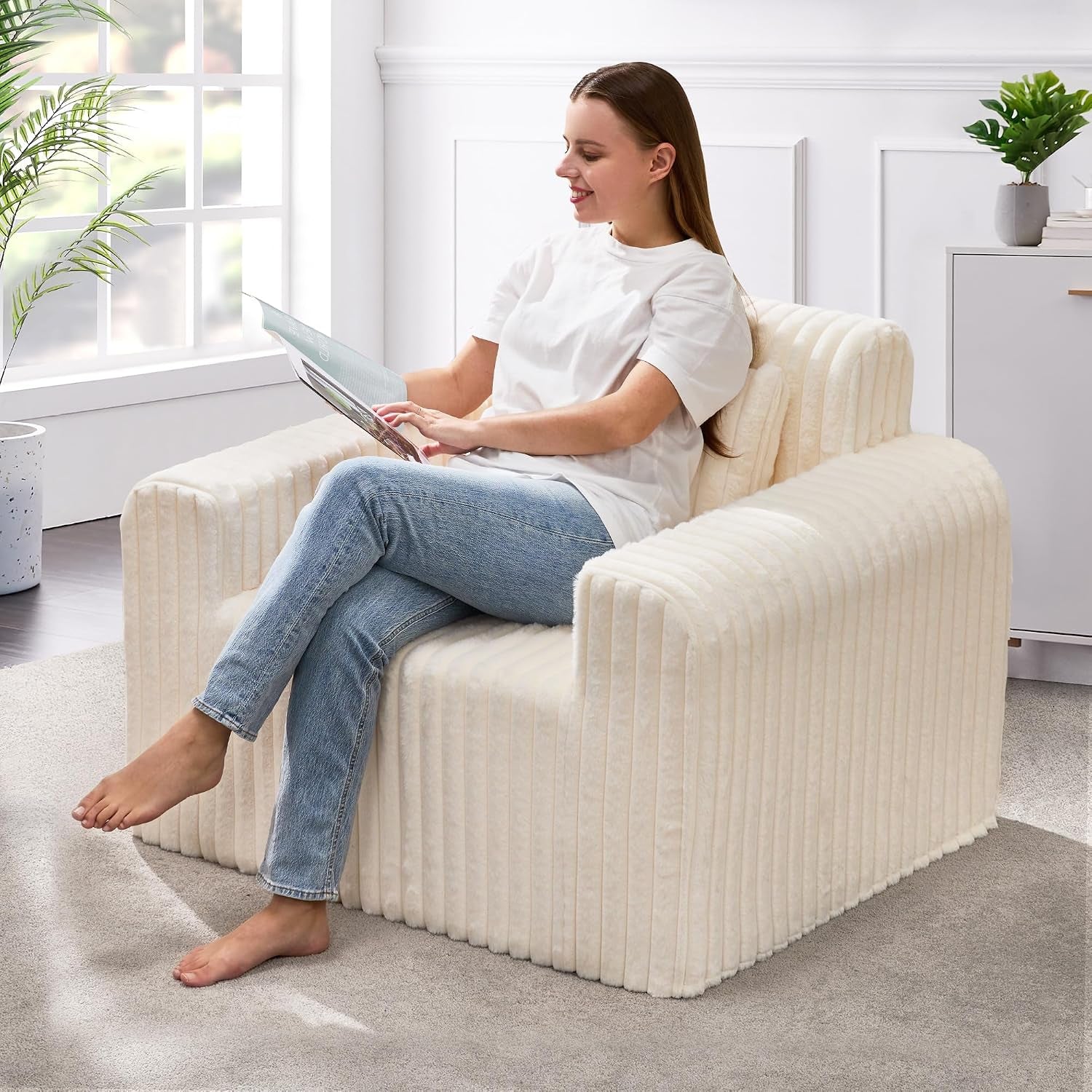 Woman reading while seated on a cream ribbed floor chair with low arms, legs crossed.