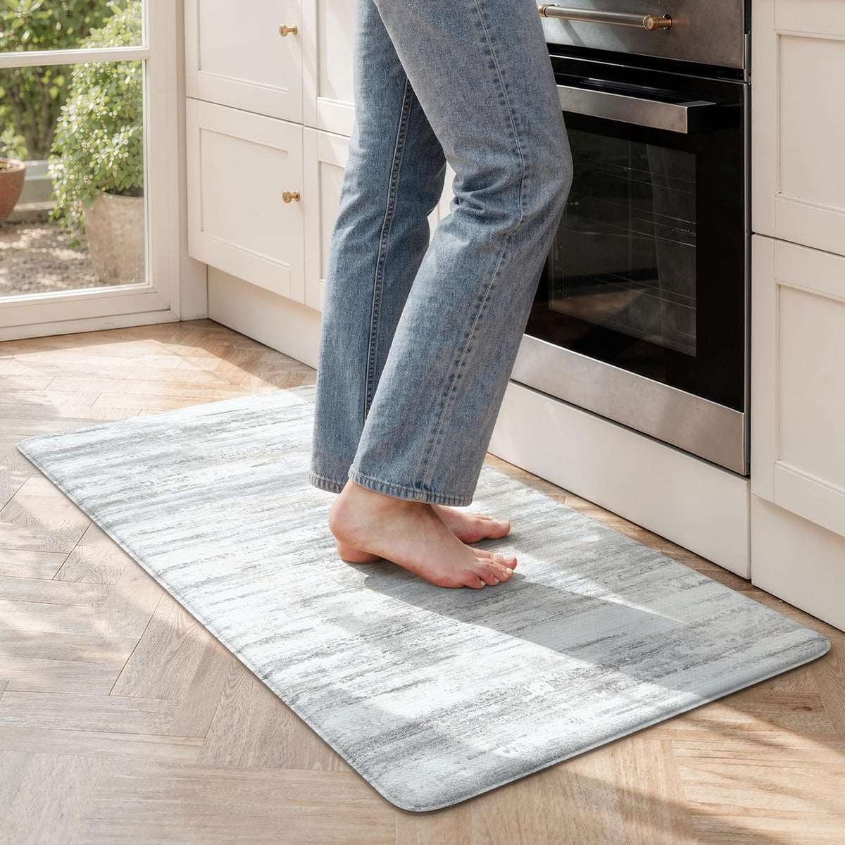 Close view of a person standing barefoot on a rectangular textured mat in front of a stainless oven on parquet flooring.