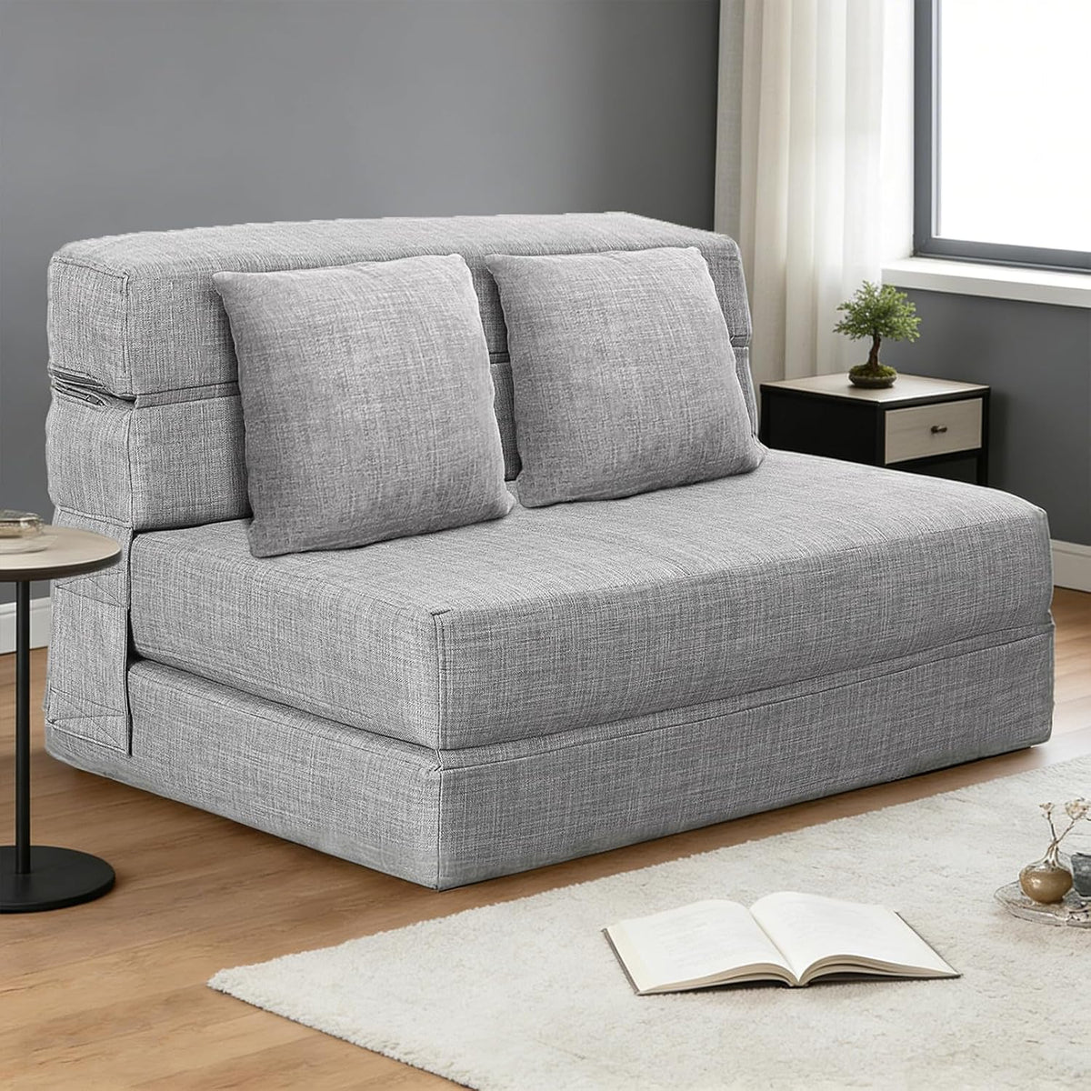 Light gray folding futon sofa in a minimal room with two matching back pillows and an open book on the rug.