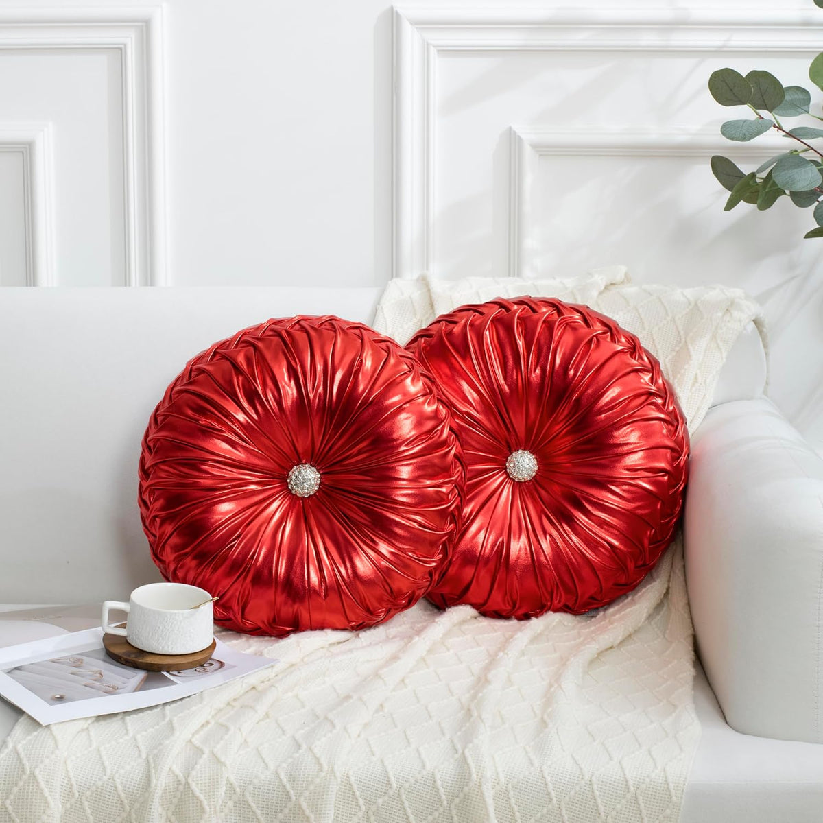 Two round ruched red satin pillows with jeweled centers on a white sofa and knit throw, tea cup nearby.