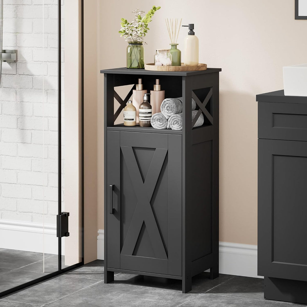 Tall narrow black floor cabinet with single door, open top shelf and decorative X sides in a tiled bathroom