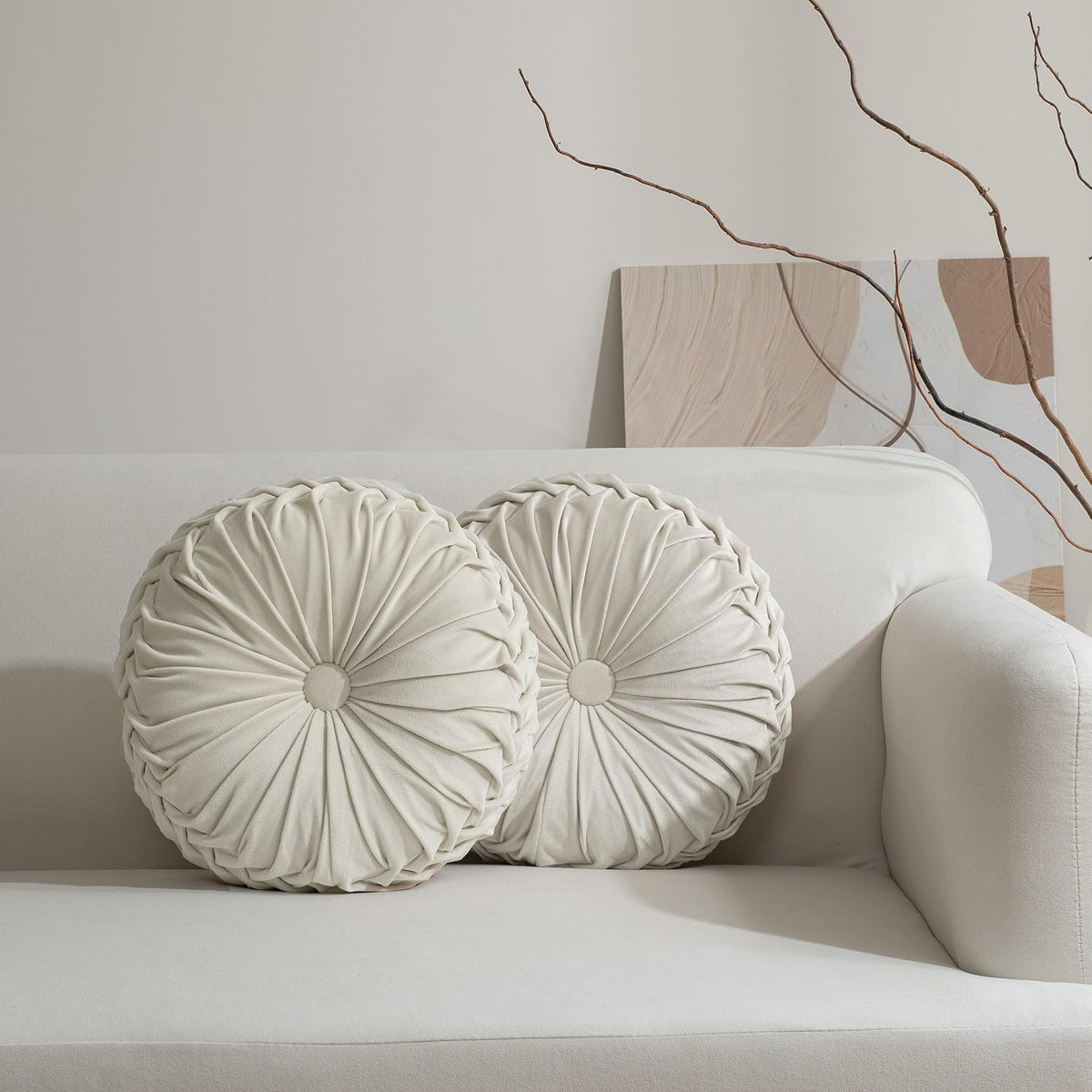 Two round ruched cream pillows with fabric button centers arranged on a light sofa.