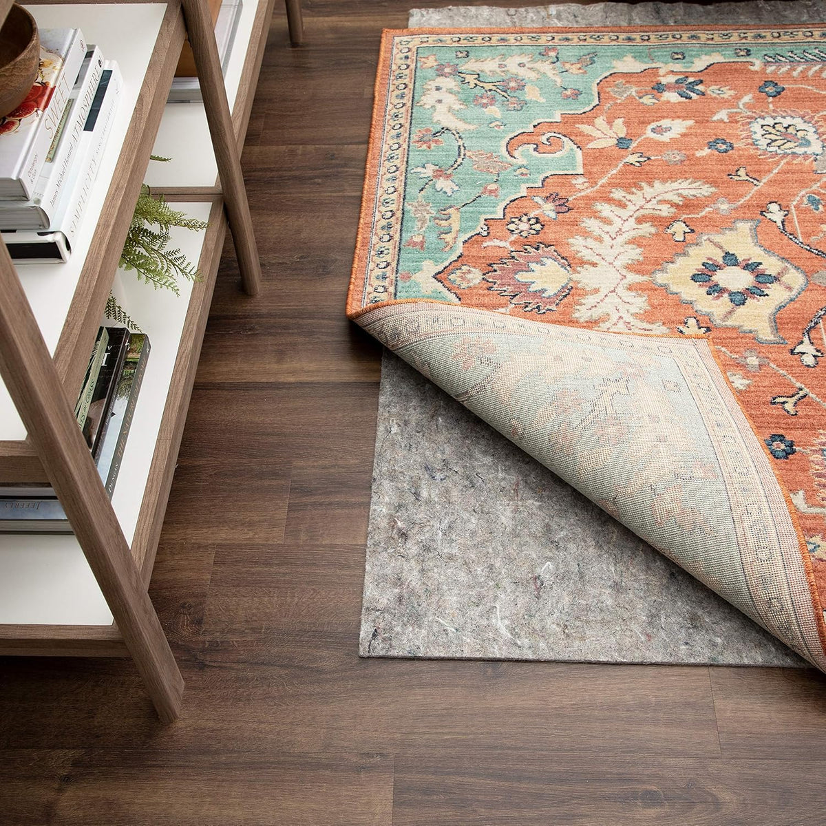 Area rug folded back at a corner to reveal a gray felt rug pad on dark hardwood next to a wooden shelf.