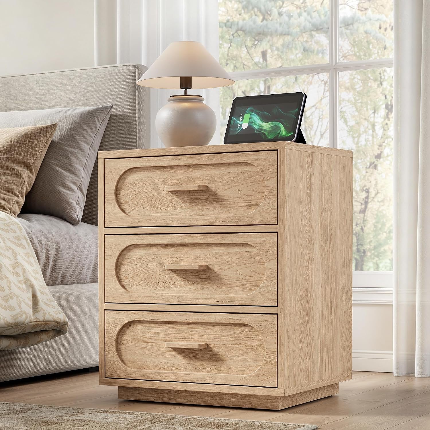Light oak three-drawer nightstand next to bed with a lamp and tablet on top, window in background.