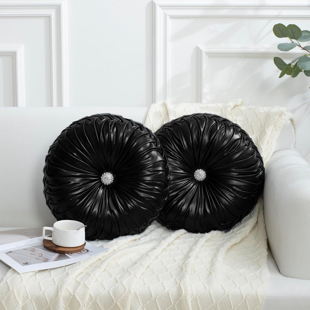 Pair of shiny black ruched round pillows with jeweled center buttons on a white sofa.