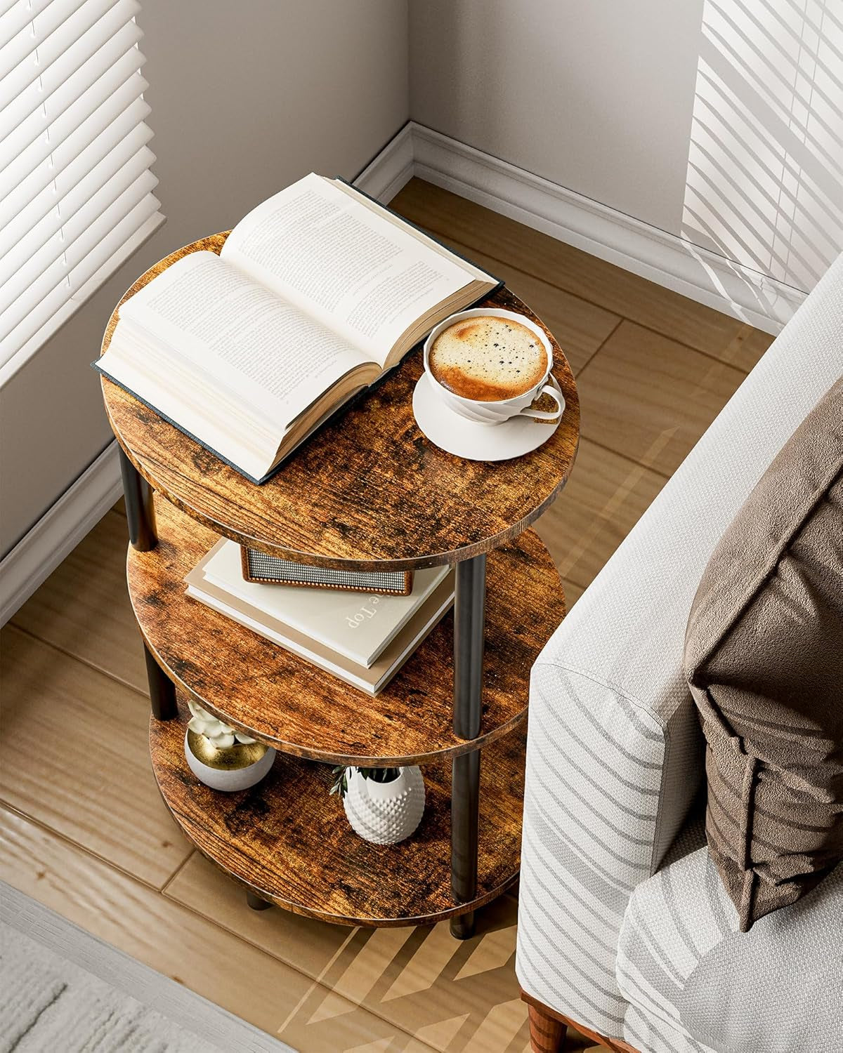Round three-tier end table staged beside a sofa with an open book and a cup of coffee on the top shelf
