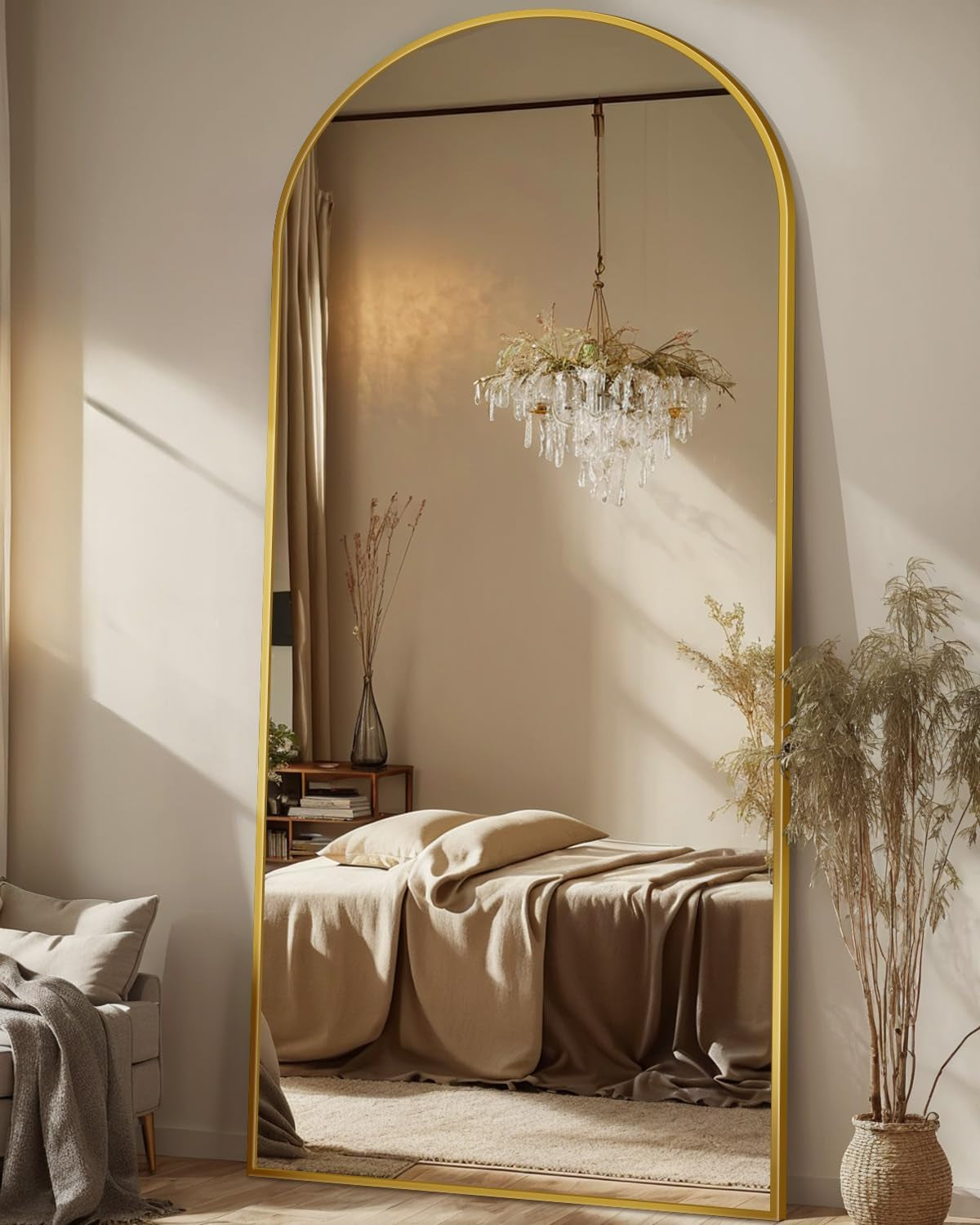 Arched full-length mirror with gold frame leaning against a neutral bedroom wall reflecting a bed and chandelier