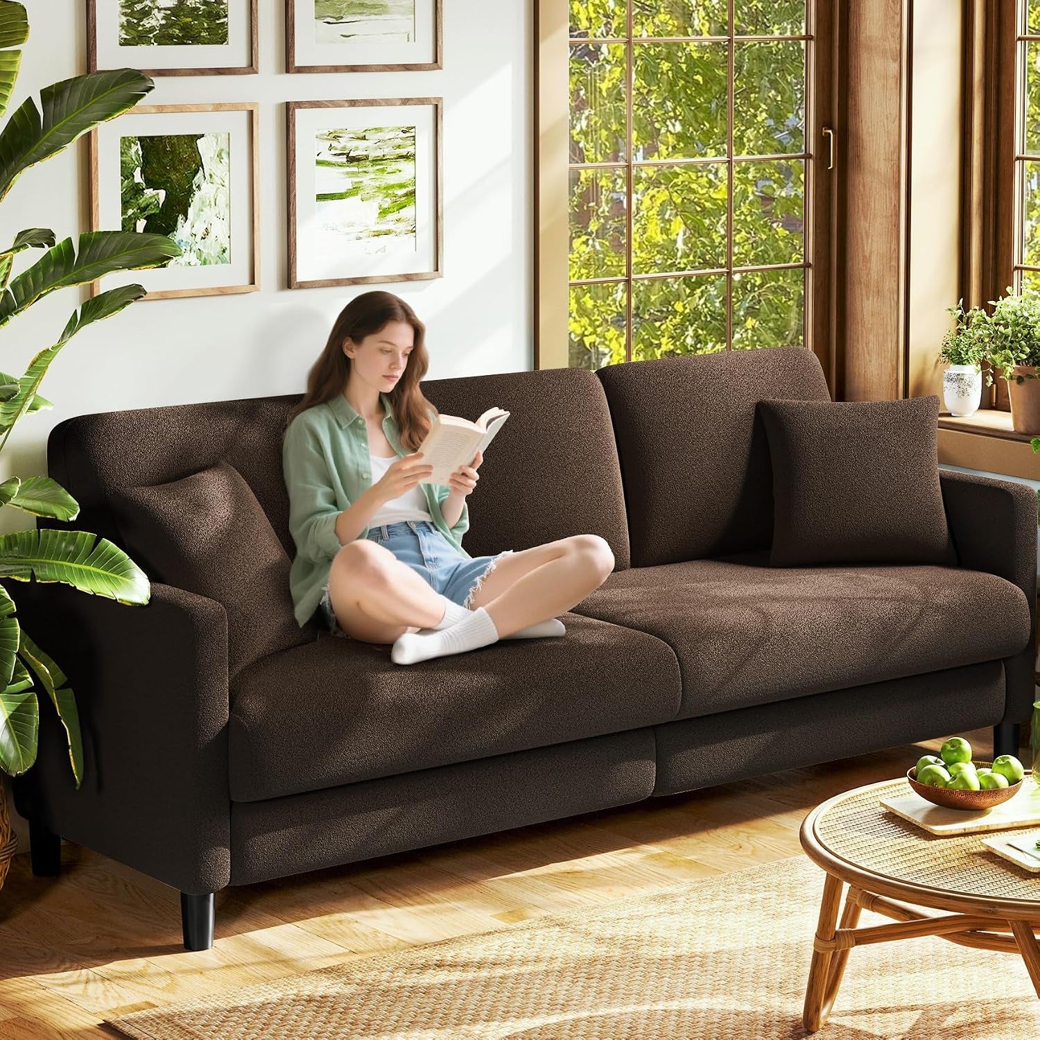 Living room scene with a brown three-seat sofa, two square pillows and a woman sitting cross‑legged reading.