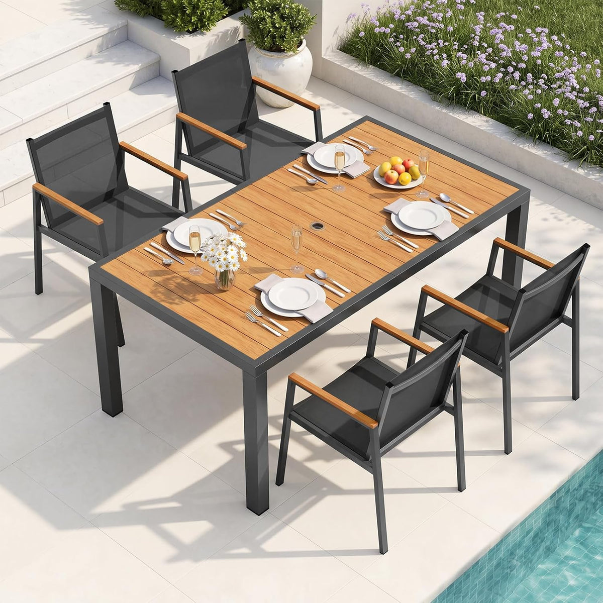 Top-down view of a dark gray-framed dining table with wood-look top and six black mesh chairs near flowerbed.