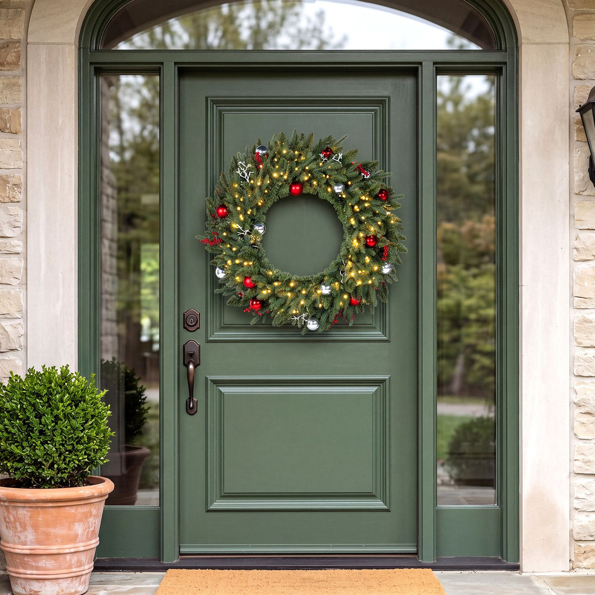 Lit ornamented evergreen wreath hanging on a dark green front door beneath an arched entryway.