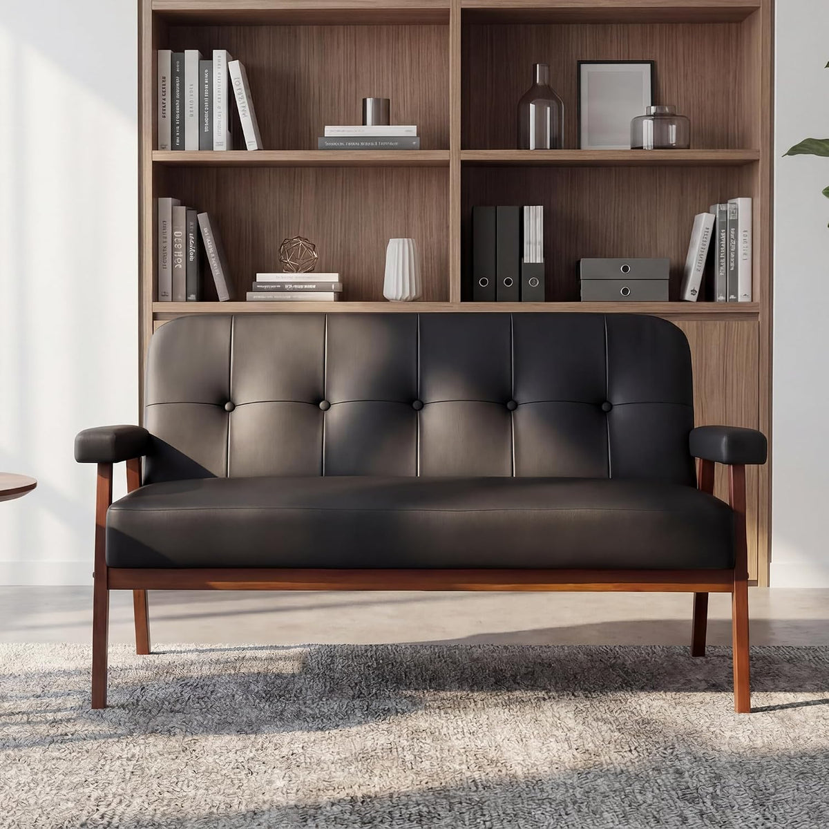 Front-facing studio view of the black tufted loveseat with walnut-colored angled legs against a bookshelf backdrop