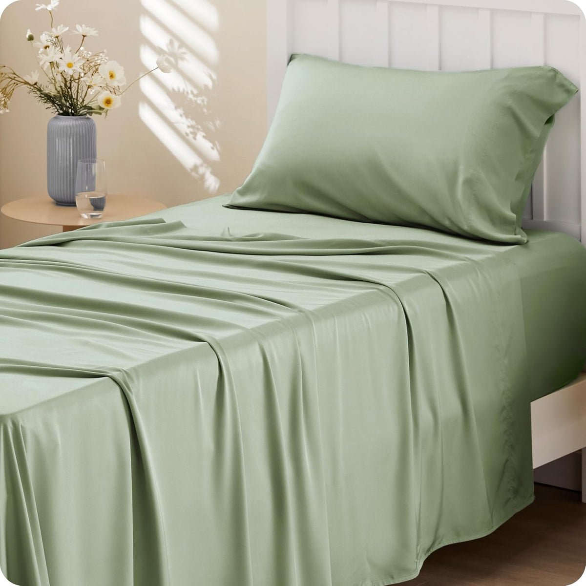 Three-quarter shot of a bed dressed in soft green sheets with gentle folds and a vase casting striped light on the wall.