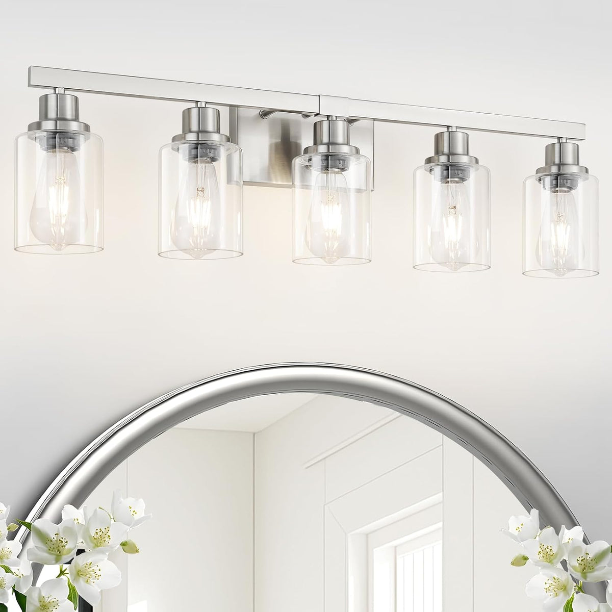 Close crop of a four-light vanity fixture in brushed nickel with clear cylindrical glass shades above a round mirror.