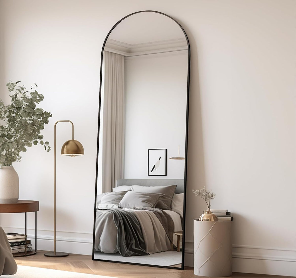 Arched full-length mirror with a thin black frame leaning against a bedroom wall, reflecting a bed.