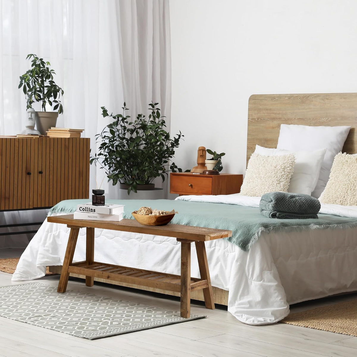Reclaimed wood bench placed at the foot of a bed in a bright bedroom with a folded teal throw, books and a wooden bowl on
