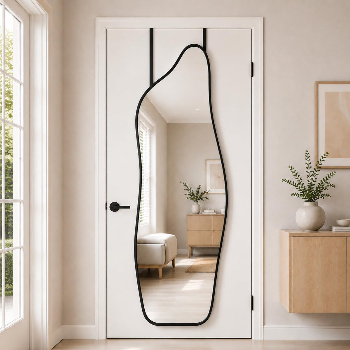 Irregular full-length mirror with a black frame hung on a door, reflecting a small bench and cabinet.