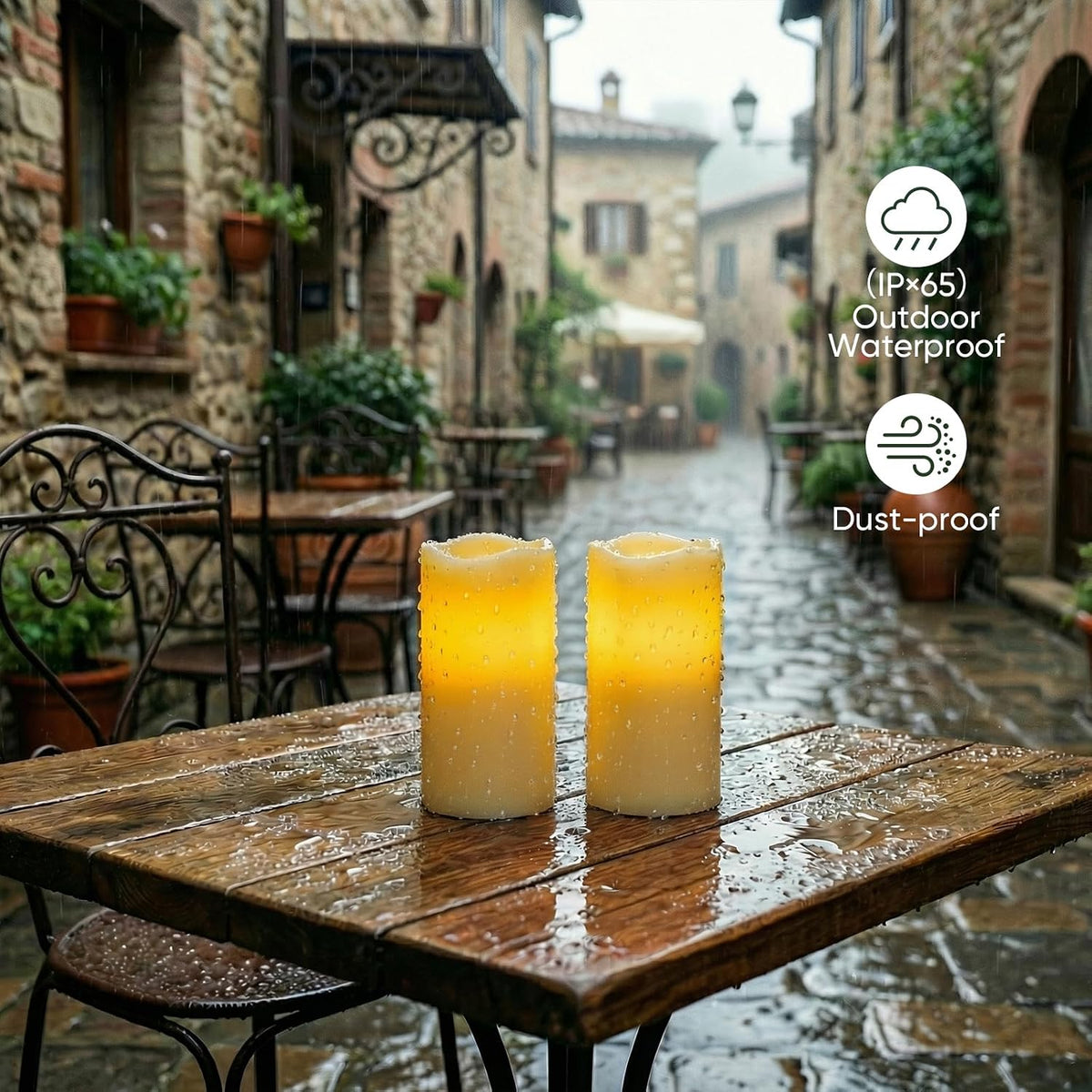 Two lit ivory resin pillar candles with water droplets on their surfaces on a rain-wet outdoor table