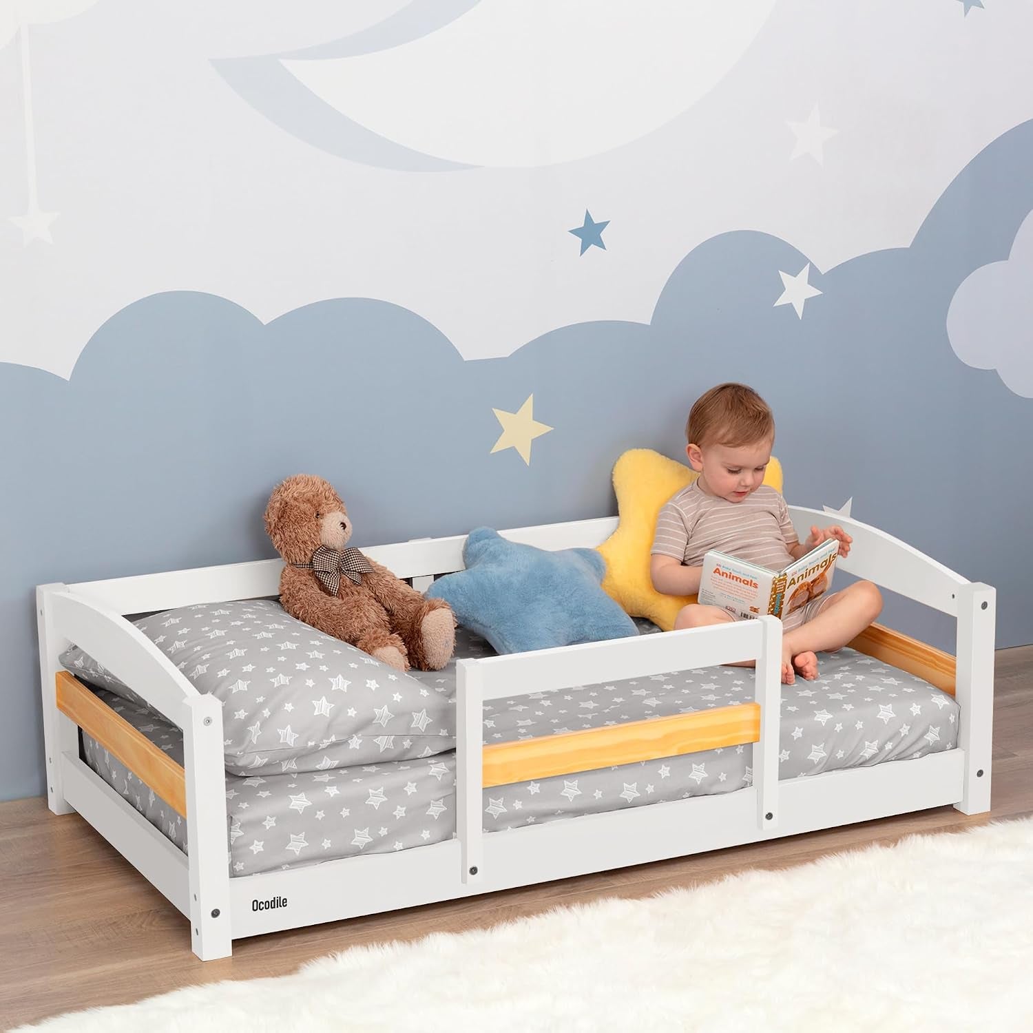 Child sitting and reading in a low white wooden toddler bed with star pillows and a teddy bear on gray star sheets.