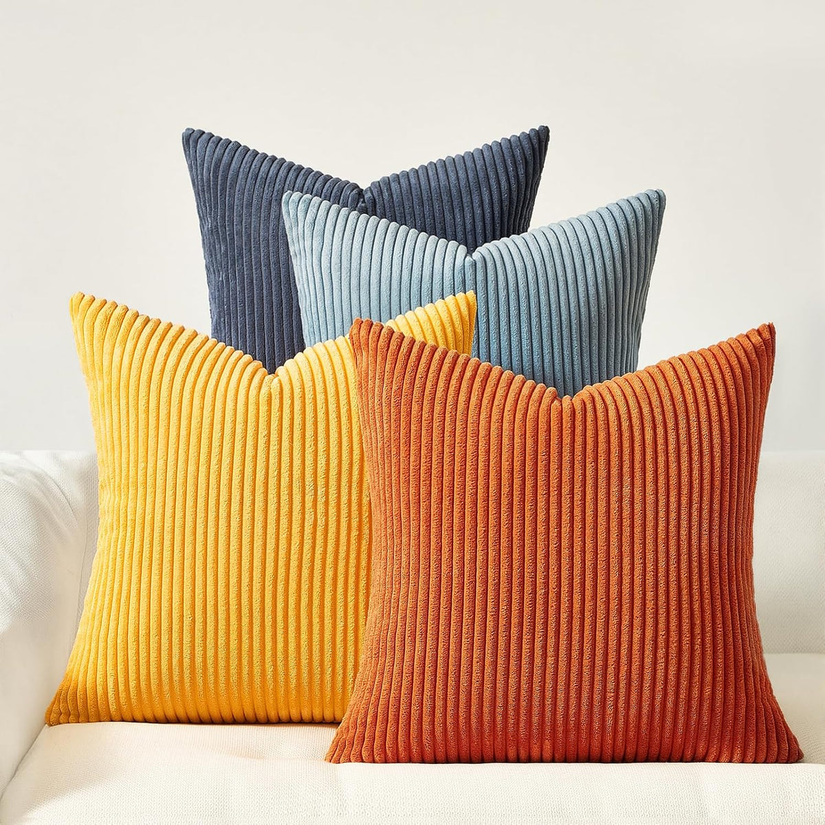 Four corduroy ribbed square pillows in burnt orange, mustard yellow, light blue and deep navy on a white sofa.