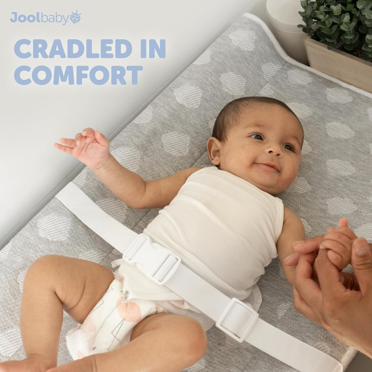 Infant lying on a grey patterned changing pad with a white safety strap while hands hold the baby
