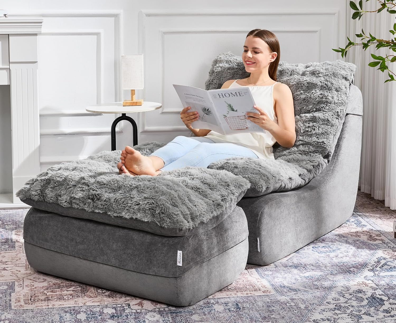 Woman reclines on a light gray plush chaise with matching ottoman and furry cushion while reading.